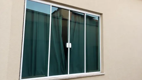 Janela de correr com quatro folhas de vidro verde, perfis brancos e puxadores centrais em parede bege clara.