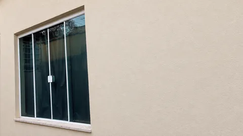 Janela de correr com quatro folhas de vidro verde, perfis prateados e peitoril de pedra em parede bege clara.