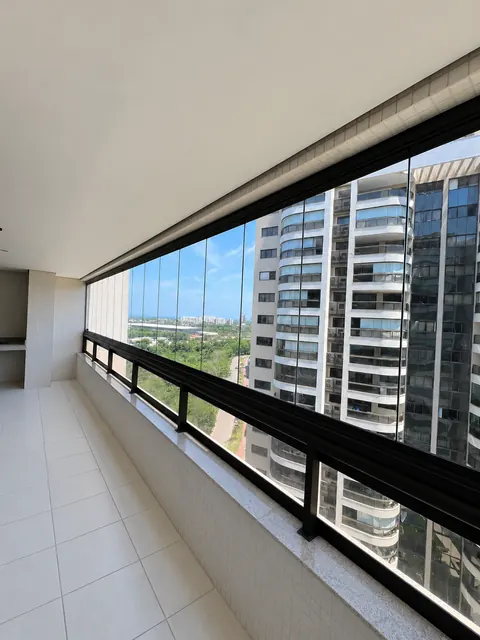 Varanda com fechamento de vidro transparente, perfis pretos e guarda-corpo, com piso limpo e vista para prédios.