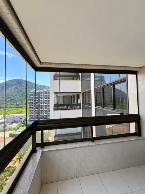 Varanda com fechamento de vidro transparente, perfis escuros e guarda-corpo, com vista para prédios e montanhas.