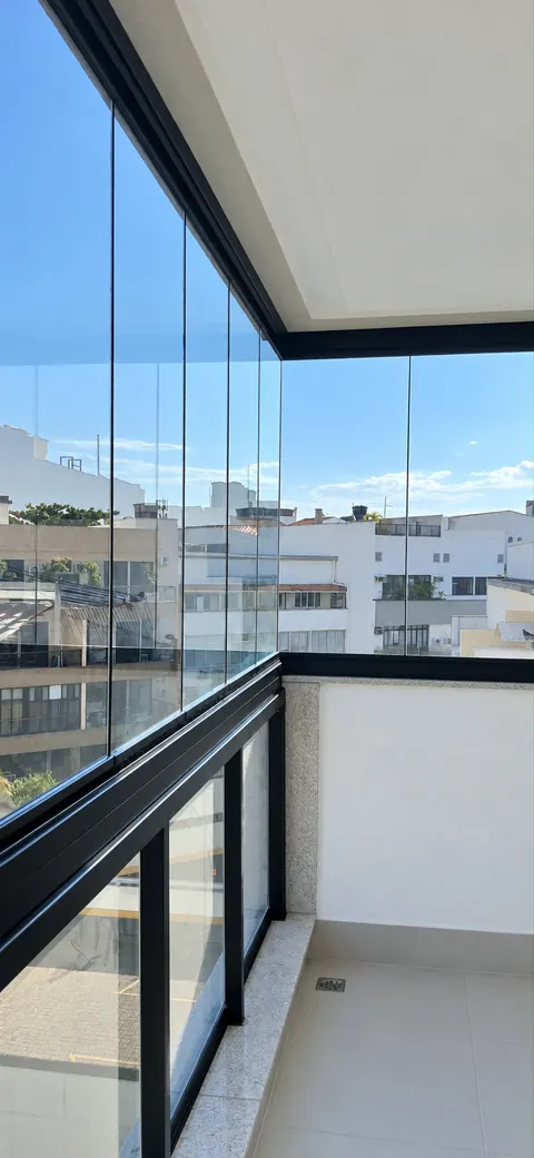 Varanda com fechamento de vidro transparente, perfis pretos e guarda-corpo envidraçado, com vista para prédios.