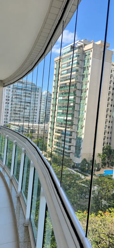 Varanda curva com fechamento de vidro e guarda-corpo branco, com vista para prédios e jardins.