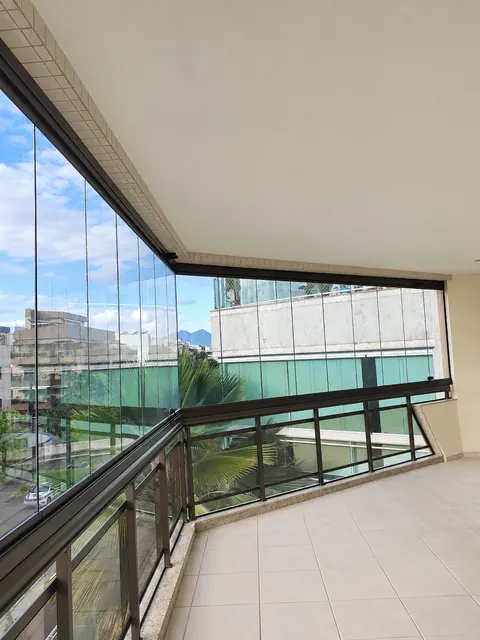 Sacada com fechamento de vidro transparente e guarda-corpo com perfis escuros, vista para prédios e palmeiras.